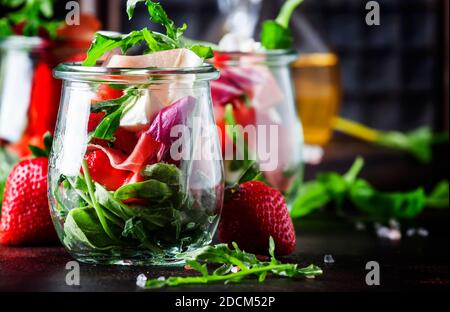 Fresh salad with spicy arugula, goat cheese, strawberry and prosciutto or ham in modern glass jars on black table, selective focus Stock Photo