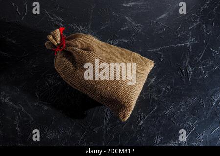 A filled Christmas bag made of coarse burlap, tied with red twine on a dark background, space for text. Copy space. Stock Photo