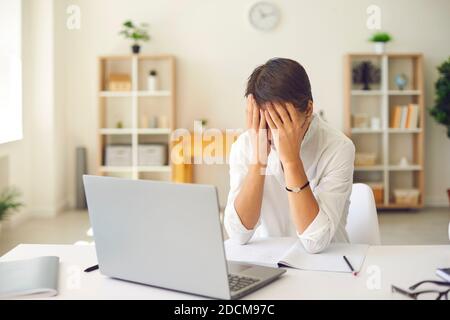Overworked female manager closes face with tired hands sitting in front of laptop in bright office. Stock Photo