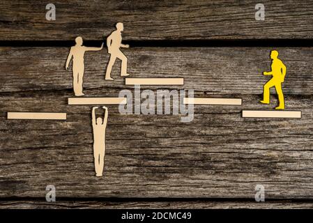 Small paper figure holding up white block as others walk on the block toward yellow paper person over wooden background. Stock Photo