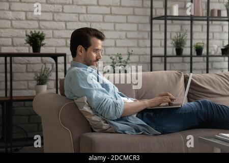 Relaxed young man lying on sofa with computer on laps. Stock Photo