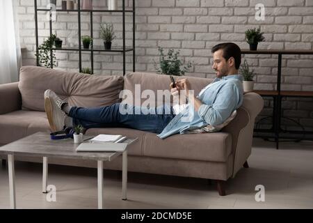 Young 30s man using mobile software application, lying on sofa. Stock Photo
