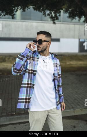 A vertical portrait of a handsome fashionable man in a check shirt and stylish sunglasses posing outdoors Stock Photo