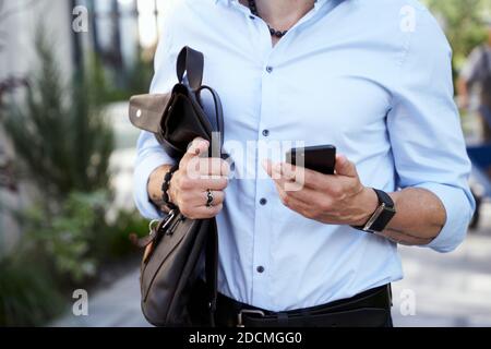 Cropped shot of modern middle aged businessman with a backpack using his smartphone while walking outdoors on a daytime Stock Photo