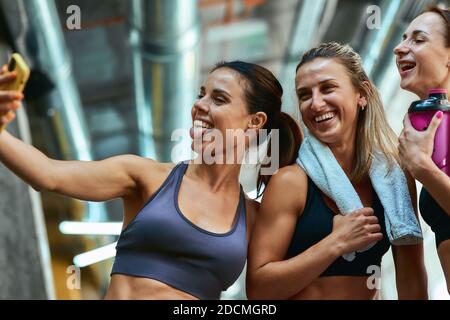 Having fun. Group of three young beautiful and joyful sportive women taking selfie on smartphone while resting after workout at gym. Sport, wellness and healthy lifestyle Stock Photo