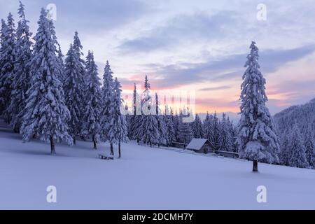Amazing winter scenery. Old wooden hut on the lawn covered with snow. Fantastic sunrise. Landscape of high mountains and forests. Wallpaper background Stock Photo