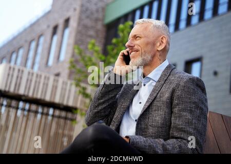 Portrait of elegant mature businessman talking on the phone while taking a break, sitting on the bench outdoors Stock Photo