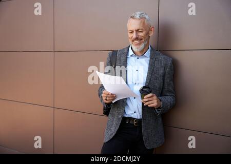 Studying information. Confident, stylish middle aged businessman smiling while working with papers, standing outdoors with a cup of coffee Stock Photo