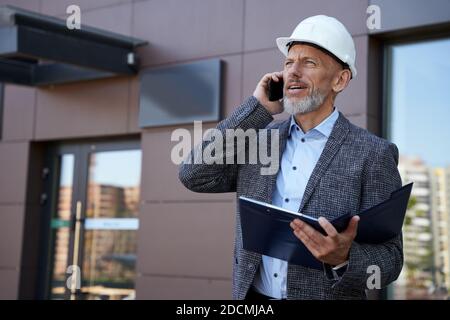 Finding solutions. Serious mature architect, engineer manager wearing helmet looking away, talking on the phone while holding a blueprint, standing Stock Photo