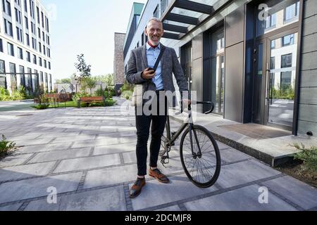 Full length shot of stylish mature businessman smiling while using his mobile phone, standing with bicycle near modern office building outdoors Stock Photo