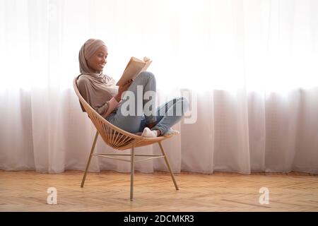 Black Muslim woman in traditional headscarf relaxing in comfy armchair with book at home, empty space Stock Photo