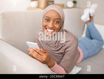 Online life. Smiling black woman in hijab lying on sofa with smartphone, chatting or browsing web at home Stock Photo