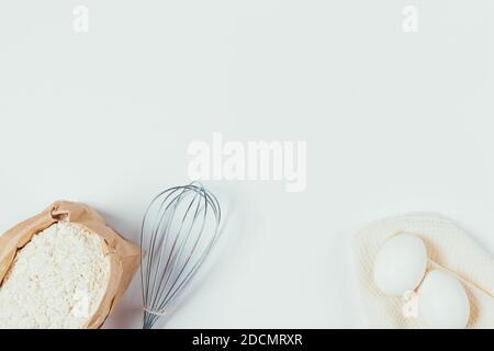 Basic ingredients for baking, paper bag with of flour and eggs next to whisk on white table, top view. Stock Photo
