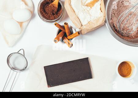 Flat lay arrangement with bar of dark chocolate next to ingredients for baking homemade brownie on white table, top view. Stock Photo
