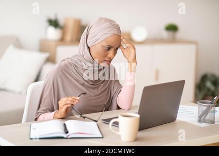 Tired black woman in hijab failing to meet deadline, having difficulty with business project near laptop at home Stock Photo
