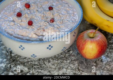 Close-up of fresh yogurt, cereals and sliced fruits, banana, apple, decorated with little strawberry from the woodland. Prepared Swiss bircher muesli. Stock Photo