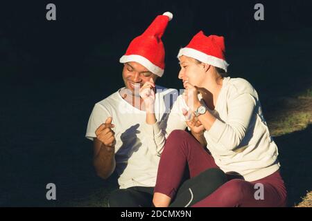 Young couple red hats singing Christmas carols sitting on floor on strong sunlight outdoors. Brazilian man and woman enjoying winter holidays Stock Photo