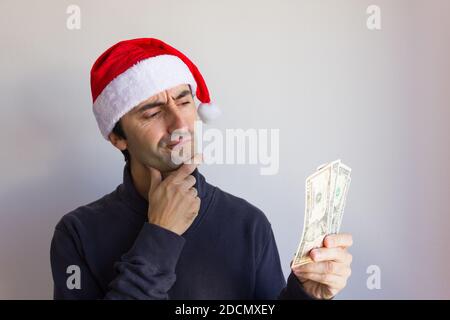 Pensive man with Santa red hat holding dollar bills over white background. Young person looking at cash with intriguing expression Stock Photo