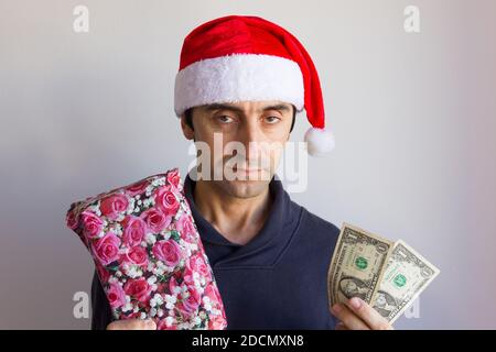 Bored young man with santa hat holding wrapped gift and little cash with disappointed face over white background. Christmas holiday season shopping Stock Photo