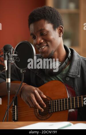 Vertical portrait of talented African-American man singing to microphone and playing guitar while recording music in studio Stock Photo