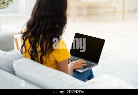 Unrecognizable black lady typing on laptop with blank screen on comfy couch at home, mockup for design Stock Photo