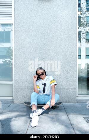 A vertical portrait of a stylish Hispanic woman in sunglasses sitting on a skateboard with an urban building behind Stock Photo