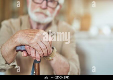 Cropped photo of senior bearded man resting at home and holding wooden walking stick. Focus on cane. Care and health concept Stock Photo