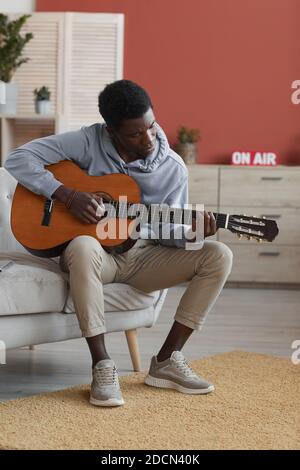 Vertical full length portrait of young African-American man playing acoustic guitar while sitting on couch at home Stock Photo