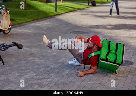 Young upset male courier in red uniform with thermo bag fell off his bicycle while riding city street. Food delivery concept Stock Photo