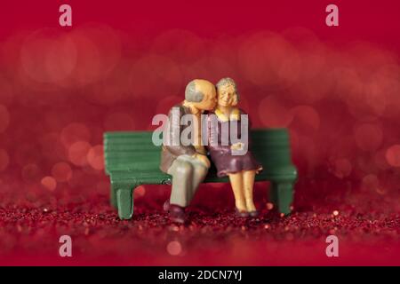 A closeup shot of a couple of old figurines sitting on a bench on red glitter background