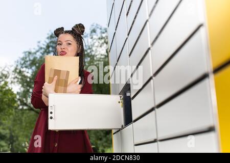 The naughty teenager shows her tongue and holds a gray envelope in her hands, which she just took out of the parcel locker. African braids. Stock Photo