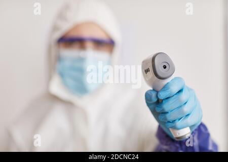 Close up of medical worker wearing full protective gear and pointing contsctless thermometer at camera while taking temperature, focus on foreground, copy space Stock Photo