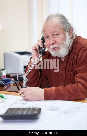 Calm senior manager calling by telephone in office room, white beard and hair, looking at camera, one person Stock Photo