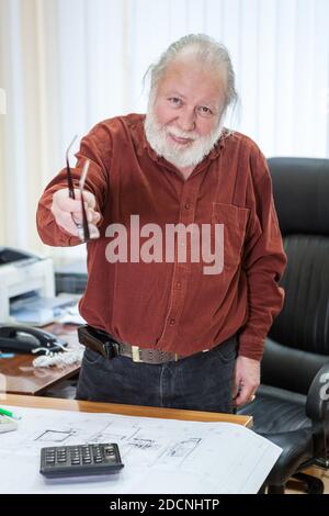 Kind and cheerful elderly male office worker points with glasses to camera Stock Photo