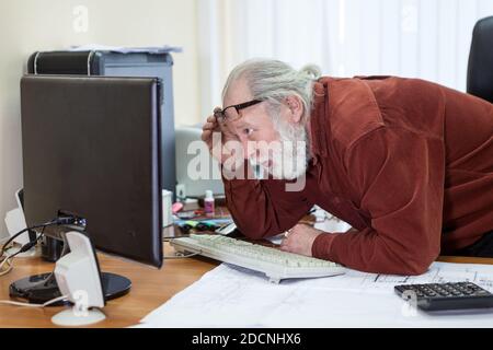 Retirement office employee looks at the monitor screen, lifting his glasses on his forehead and moving close Stock Photo