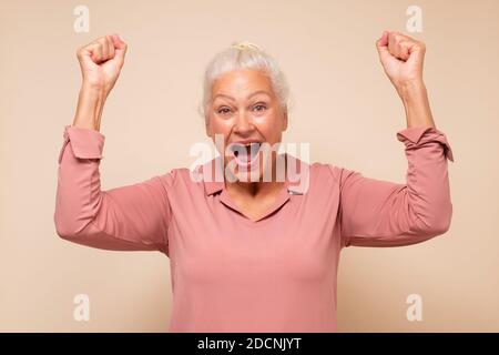 Happy middle aged caucasian woman raising fists in winning gesture and celebrating success. Mature woman hitting jackpot. Win and success concept Stock Photo