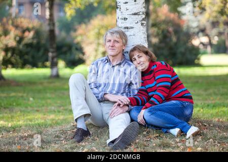 Senior Caucasian couple sitting under birch tree on earth in autumn park, holding hands and resting Stock Photo