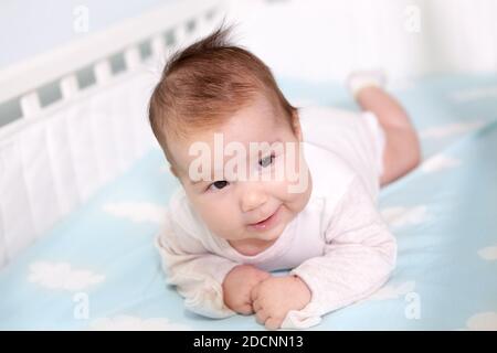 Sweet smiling newborn boy lying on his stomach in cot, trying to hold her head and looks forward Stock Photo
