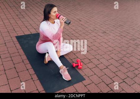 Sports girl drinks water from bottle while sitting on the yoga mat after training. Thirst female athlete Stock Photo
