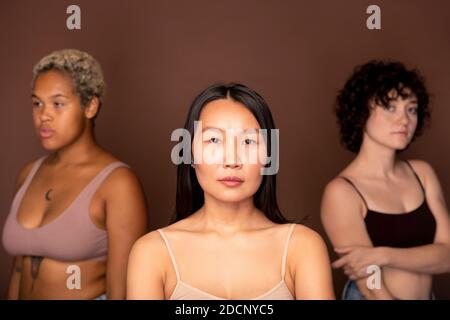 Young serious Asian woman in beige tanktop standing against two other females Stock Photo