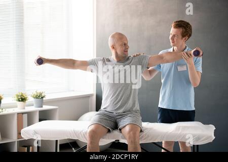 Young physiotherapist standing behind bald mature man with dumbbells in hands Stock Photo