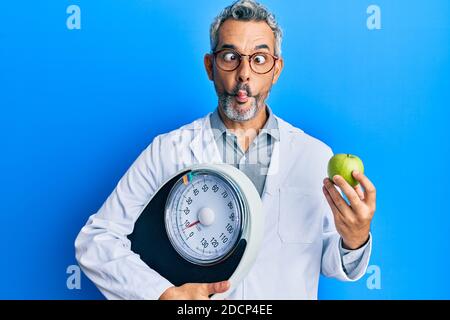 Middle age grey-haired man as nutritionist doctor holding weighing machine and green apple making fish face with mouth and squinting eyes, crazy and c Stock Photo