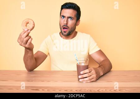 Young hispanic man sitting at the table drinking chocolate beverage holding donut in shock face, looking skeptical and sarcastic, surprised with open Stock Photo