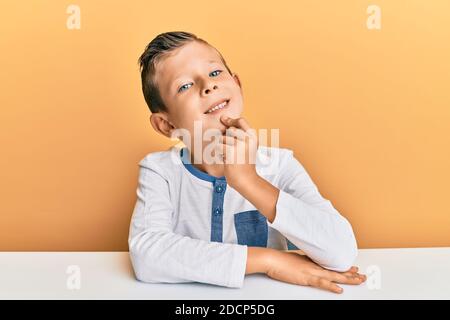 Adorable caucasian kid wearing casual clothes sitting on the table looking confident at the camera with smile with crossed arms and hand raised on chi Stock Photo