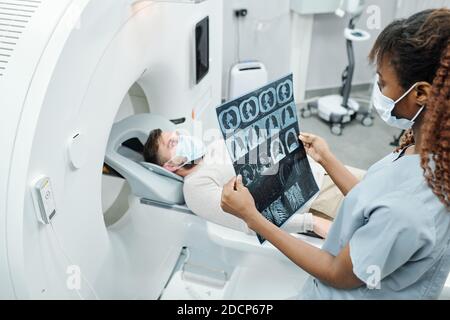 Young African radiologist in uniform and protective mask looking at x-ray image Stock Photo