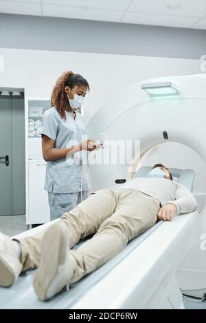 Young African female assistant in medical uniform and mask looking at patient Stock Photo