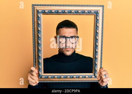 Handsome man with tattoos holding empty frame skeptic and nervous, frowning upset because of problem. negative person. Stock Photo