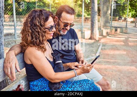 Middle age couple smiling happy using smartphone. Sitting on the bench with chihuahua dog at park Stock Photo