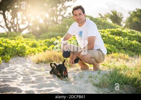 Young fit male squats on the beach with his small black dog in Hawaii Stock Photo