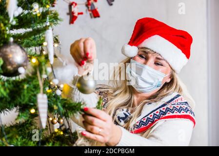 Middle age blond woman with blue eyes in Christmas hat and face protection arranging Christmas tree with decorations Stock Photo